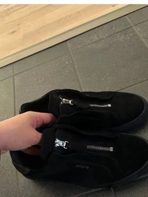 Svarta Arigato sneakers med dragkedja - Stilrena svarta sneakers från Arigato i mocka med silverfärgade dragkedjor istället för klassiska snören. Låg modell med minimalistisk design och diskret logga på sidan. Perfekta för dig som gillar en clean look och vill ha något unikt på fötterna.