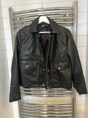 Svart skinnjacka från Zara - Snygg svart skinnjacka (konstläder) från Zara med klassisk krage, dragkedja framtill och två stora fickor tryckknappar. Jackan har en rak passform och stilrena detaljer som passar till många olika looks. Använd max 3 gånger 
