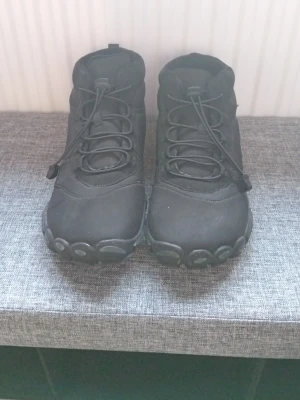 Svarta minimalistiska kängor - Säljer ett par svarta kängor med minimalistisk design och tåskydd. Skorna har snörning och är tillverkade i ett slitstarkt material, troligen syntet eller mockaimitation. Perfekta för dig som gillar en clean look och vill ha bekväma skor till vardags.