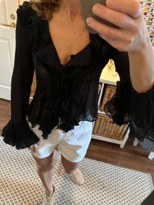 Svart blus volangblus med spetsdetaljer Stl 38  - Snygg svart blus i transparent material med volanger och spetsdetaljer längs ärmar och nederkant. Djup v-ringning och långa, vida ärmar med flera lager volanger ger en romantisk och trendig look. Perfekt för dig som gillar statement-plagg. Paris . Står Stl 3 , är Fransk Stl så den är som Stl 38