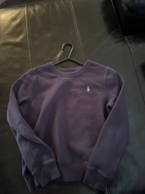 Mörkblå sweatshirt från Ralph Lauren - Mörkblå sweatshirt med rund hals från Ralph Lauren. Tröjan har en broderad logga på bröstet och ribbade muddar vid ärmslut och nederkant. Perfekt för en avslappnad och stilren look.