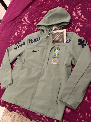 Turkos Brasil vindjacka med huva - Ljusgrå vindjacka storlek L (passar M och S)  med Brasiliens landslagslogga och flera sponsortryck. Jackan har huva, hel dragkedja och är tillverkad i GORE-TEX-material som står emot regn och vind. Perfekt för dig som gillar sportig stil och vill representera Brasilien. Oan