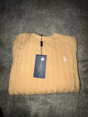 Beige  tröja Polo Ralph Lauren - Snygg beige kabelstickad tröja från Polo Ralph Lauren med rund halsringning och den klassiska broderade loggan i vitt på bröstet. Tröjan har långa ärmar och ribbade muddar vid ärmslut och nederkant. Perfekt för en stilren och avslappnad look.