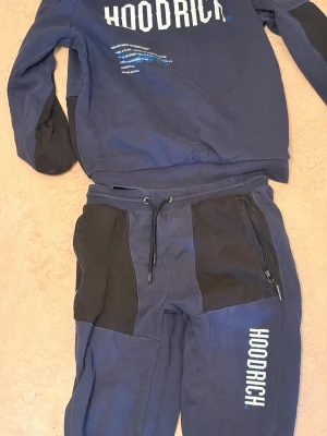 Blått Hoodrich loungewear-set - Snyggt blått loungewear-set från Hoodrich med matchande tröja och byxor. Setet har svarta detaljer på armbågar och byxfickor, samt vita Hoodrich-loggor på både tröja och byxa. Tillverkat i mjukt bomullsmaterial med dragkedjefickor och justerbar midja.