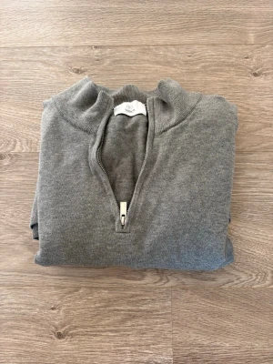 Grå halvzip-tröja från bläck - Snygg grå tröja från Bläck med halv dragkedja och hög krage. Tillverkad i mjukt material och har långa ärmar samt ribbade muddar vid ärmslut och nederkant. Perfekt för lager på lager och enkel att matcha med olika outfits.