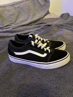 Svarta Vans Old Skool sneakers - Klassiska Vans Old Skool sneakers i svart mocka och canvas med vit sula och vit sidostripes. Skorna har vita snören och den ikoniska röda Vans-loggan på hälen. Perfekta för en avslappnad streetwear-look. Storlek 36- 22,5 cm lång fot. Ny pris 599kr mitt pris 350