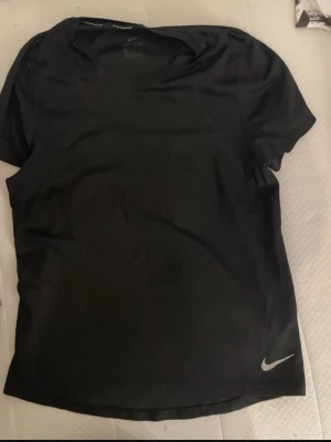 Svart Nike träningströja - Snygg svart träningströja från Nike med korta ärmar och rund hals. Tillverkad i ett lätt och ventilerande material som passar perfekt för träning. Diskret Nike-logga i vitt nere vid fållen. Enkel och sportig design.
