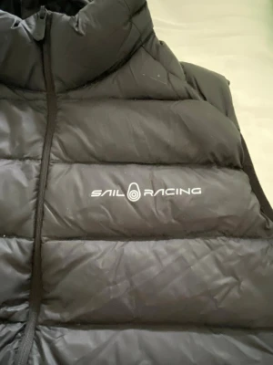 Svart dunväst från Sail Racing - Snygg svart dunväst från Sail Racing med hög krage och dragkedja framtill. Västen har ett stilrent tryck med logga på bröstet och en större logga på ryggen. Tillverkad i ett glansigt material som ger en sportig look. Perfekt för lager-på-lager under kyliga dagar. Finns inga defekter alls på varan. 
