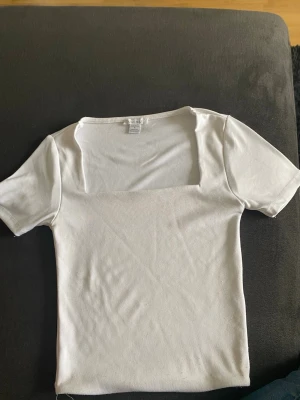Vit topp med fyrkantig urringning  - Säljer en enkel och stilren vit topp med fyrkantig urringning och korta ärmar. Toppen är tight i modellen och passar perfekt till jeans eller kjol. Materialet är mjukt och stretchigt, vilket gör den bekväm att bära hela dagen. Lite av en magtröja också.