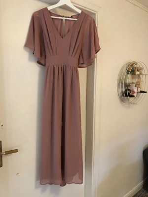 Gammelrosa långklänning med v-ringning - Säljer en elegant gammelrosa långklänning med v-ringad hals och vida, korta ärmar. Klänningen har fina veck framtill, ett knytband i midjan och är tillverkad i ett lätt, flytande material som ger ett snyggt fall.
