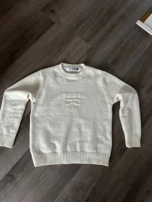 Vit stickad Prada tröja  - Säljer en stilren vit stickad tröja från Prada med broderad logga och text framtill. Tröjan har rund halsringning, ribbade muddar och nederkant. Perfekt för dig som gillar klassisk och enkel design.