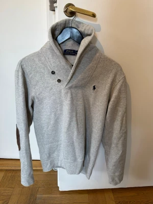 Grå tröja från Polo Ralph Lauren - Snygg grå tröja från Polo Ralph Lauren med hög krage och två knappar vid halsen. Tröjan har mörkblå logga broderad på bröstet och förstärkningar på armbågarna. Perfekt för dig som gillar klassisk och stilren design.