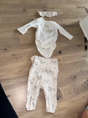 Sött babyset med svanar och hårband - Sött babyset med svanar och hårband Tre-delat babyset i mjuk bomull, består av en långärmad body i offwhite med svanmotiv, matchande byxor med svanprint och ett gulligt hårband. Perfekt för dig som gillar söta och bekväma outfits till de minsta.