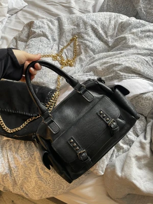 Svart handväska med nitar och spännen - Snygg svart handväska i skinnimitation med dubbla handtag. Väskan har dekorativa nitar och spännen på framsidan samt en praktisk ficka. Klassisk boxig form som rymmer det mesta du behöver. Perfekt accessoar för en trendig look.