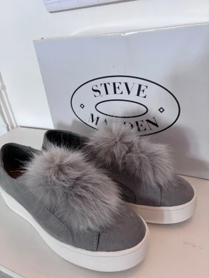 Grå loafers med fuskpäls Steve Madden - Grå loafers från Steve Madden med bred vit platåsula och stor fluffig detalj i grå fuskpäls på ovansidan. Skorna har en stilren slip-on design och är tillverkade i textil och syntetmaterial. Perfekta för dig som vill sticka ut med din stil. Storleken är snarare en 39.
