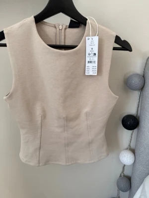 Beige ärmlös topp från Gina tricot - Snygg beige ärmlös topp från Gina tricot med figurnära passform och rund halsringning. Toppen har diskreta sömdetaljer framtill och dragkedja i ryggen. Perfekt att styla med jeans eller kjol för en trendig look. Helt oanvänd, prislapp är kvar 