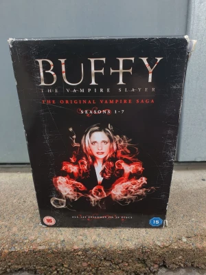Dvd - Box med Buffy The Vampire Slayer: The Original Vampire Saga, säsong 1-7. Svart kartong med röd och vit grafik och text. Innehåller alla avsnitt. Perfekt för dig som gillar vampyrserier och klassiska tv-shower.
