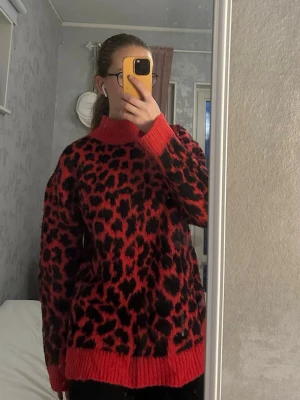 Röd leopardmönstrad stickad tröja - Cool stickad tröja i rött med svart leopardmönster. Tröjan har hög krage, långa ärmar och ribbade muddar vid ärmslut och nederkant. Perfekt för dig som vill sticka ut med en trendig och djärv look. I verkligheten är den mer mörkrosa än röd.