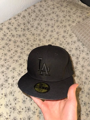 New Era keps - Snygg svart keps från New Era med LA-logga broderad framtill. Modellen är 59FIFTY med rak skärm och klassisk passform. Undersidan av skärmen är ljusgrå och insidan har detaljerade band med 59FIFTY-tryck. Perfekt för dig som gillar streetwear och sportig stil. Storlek 7 1/8 aldrig använd