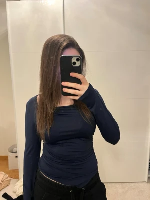 Mörkblå långärmad topp från H&M - Snygg mörkblå långärmad topp från H&M med figurnära passform och lätt rynkade detaljer på sidan. Toppen har en bred, något asymmetrisk ringning och är tillverkad i ett stretchigt syntetmaterial som sitter bekvämt på kroppen.