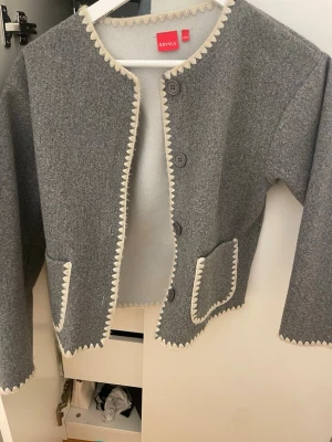Grå cardiganjacka från ryvls  - PRIS KAN DISKUTERAS. Super fin grå cardiganjacka med dekorativa vita kanter och stora knappar framtill. Jackan har två framfickor och är tillverkad i ett tjockare ull-liknande material. Rak passform och långärmad, perfekt för våren. Originalpriset var 600kr jag är 162 cm och den passar 