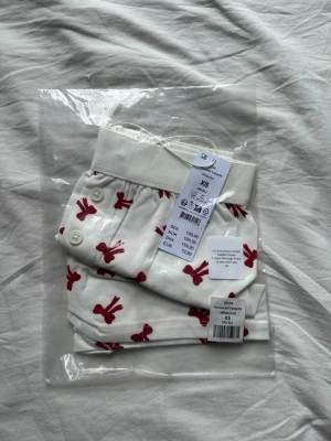 Vita trosor med röda rosetter  - Vita pjamas shorts från Gina Tricot med sött mönster av röda rosetter. Modellen har bred resår i midjan och dekorativa knappar på sidan. Tillverkade i mjuk bomull för skön känsla hela dagen.