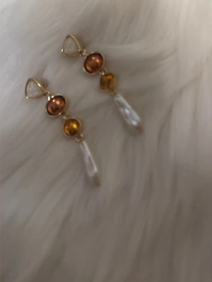 Guldiga örhängen med pärlor och stenar - Unika örhängen i guldton med droppformade vita pärlor, orange och gula stenar samt en triangelformad klar sten upptill. Perfekta för dig som gillar färg och statement-accessoarer. Säljs som par.