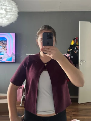 Vinröd kortärmad kofta med knapp - Snygg vinröd kofta med korta ärmar och en guldig knapp upptill. Koftan har öppen framsida och rak passform, perfekt att slänga över ett linne eller t-shirt. Materialet är mjukt och stretchigt, vilket gör den bekväm att bära.