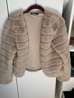 Beige pälsjacka från Gina Tricot - Supersnygg beige pälsjacka från Gina Tricot i fuskpäls. Jackan har en fluffig och mjuk struktur med horisontella sömmar som ger en cool look. Den är öppen framtill och har långa ärmar. Perfekt för att hålla sig varm och trendig under vintern.