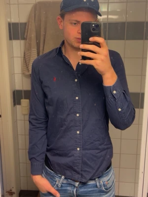 Marinblå skjorta från Ralph Lauren - Klassisk marinblå skjorta från Ralph Lauren med knappstängning och den ikoniska röda logotypen broderad på bröstet. Skjortan har lång ärm, normal passform och är tillverkad i mjuk bomull. Perfekt till jeans eller chinos för en clean look.