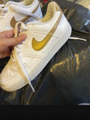 Nike vita sneakers med guld swoosh - Snygga vita sneakers från Nike med metallic guld swoosh och detaljer på hälen. Klassisk låg modell med perforerad tå och vita skosnören. Tillverkade i läder för en clean och stilren look som sticker ut.