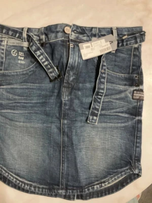Blå jeanskjol från G-Star RAW - Säljer en snygg blå jeanskjol från G-Star RAW med klassiska fem fickor, bälteshällor och en cool tvättad denimlook. Kjolen har en kort längd och raka sömmar, samt detaljer som knapp och dragkedja framtill. Perfekt för dig som gillar streetstyle.