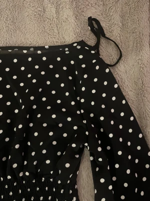 Svart prickig topp - Supersöt svart klänning med vita prickar och långa ärmar. Klänningen har knytband på axlarna och smock i midjan för en bekväm passform. Perfekt för dig som gillar en lekfull och trendig stil.