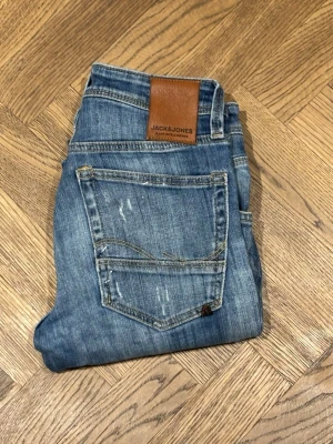 Blå slitna jeans från Jack & Jones - Säljer ett par blå jeans från Jack & Jones med slitningar och hål på benen för en trendig look. Jeansen har klassisk femficksmodell, normal midja och raka ben. Tillverkade i bomull med en snygg tvättad finish och läderpatch bak i midjan. Storlek 27/32 sitter större i midjan 