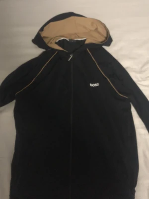 Svart vindjacka med huva från BOSS - Svart vindjacka från BOSS med dragkedja och huva. Jackan har beige innerfoder i huvan och tunna beige detaljer längs axlarna. BOSS-logga broderad på bröstet. Perfekt för blåsiga dagar och enkel att matcha med olika outfits.