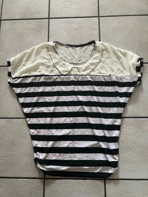Randig topp storlek L - Snygg topp med svarta och vita ränder samt en dekorativ pärlkrage. Överdelen är vit och har en lös passform med korta ärmar. Perfekt för dig som gillar unika detaljer och en lekfull stil.