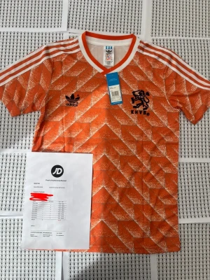 Holland retro Van Basten fotbollströja Adidas - Säljer en ikonisk orange Holland retro fotbollströja från Adidas med vita och svarta detaljer. Tröjan har klassiskt mönster, vita ränder på axlarna, svart KNVB-logga och Adidas-märke fram. På ryggen står det VAN BASTEN och nummer 12 i svart. Perfekt för fotbollsfans!