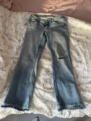 Ljusblå bootcut jeans med hål - Säljer ett par ljusblå bootcut jeans med slitning och hål på ena benet. Jeansen har klassisk femficksmodell och råa kanter vid bensluten. Perfekta för en avslappnad och trendig look. Köpta från lindex men har gjort hålen och sprättat upp benen. Lite korta på mig som är 176. Kan tänka mig att de passar nån med storlek 36 också om man vill ha en lite lösare look.