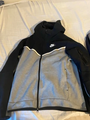 Nike hoodie med dragkedja svart/grå - Säljer en snygg hoodie från Nike med dragkedja. Den är svart upptill och på ärmarna, grå på nederdelen och har en vit detalj längs bröstet. Nike-loggan är broderad på bröstet. Hoodien har långärmad design och är tillverkad i mjuk bomull och polyester.