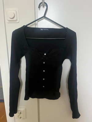 Svart ribbad topp med spets från Zara - Svart långärmad topp från Zara med ribbad struktur och dekorativa spetsdetaljer vid bysten. Toppen har v-ringad urringning och knappar framtill som ger en snygg touch. Perfekt att styla med jeans eller kjol för en trendig look.