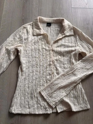 Vit plisserad blus med krage - Trendig vit plisserad blus från Monki med lång ärm och klassisk krage. Materialet är lätt och har en snygg struktur som ger en cool look. Perfekt att styla med jeans eller kjol för en modern outfit.