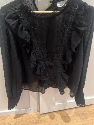 Svart volangblus  - Snygg svart blus  med volangdetaljer framtill och prickig meshstruktur. Blusen har långa ärmar och spetsdetaljer längs kanterna. Använd 1 gång.