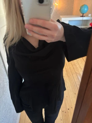 Svart långärmad blus med draperad krage - Stilren svart blus med lång ärm och snyggt draperad krage. Blusen har en figurnära passform och är tillverkad i ett mjukt, följsamt tyg som ger en elegant look. Perfekt att matcha med både jeans och kjol för en trendig outfit.