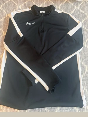 Svart Nike träningströja med dragkedja - Snygg svart långärmad träningströja från Nike med vit logga på bröstet och vita ränder längs ärmarna. Tröjan har en halv dragkedja framtill och står för sportig stil, perfekt för träning eller uppvärmning.