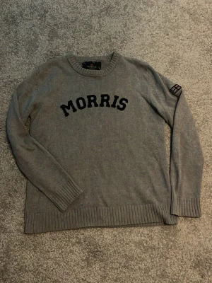 Grå stickad tröja från Morris - Snygg grå stickad tröja från Morris med svart text på bröstet och en patch med brittiska flaggan på ärmen. Tröjan har rund halsringning, långa ärmar och ribbade muddar. Perfekt för en avslappnad men stilren look.