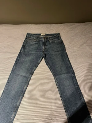 Blå slim straight jeans Jack & Jones - Säljer ett par blå slim straight jeans från Jack & Jones, modell Tim. Klassisk femficksdesign, normal midja och raka ben. Jeansen är i slitstark denim med snygga kontrastsömmar och metallknappar. Perfekta för en avslappnad och trendig look.
