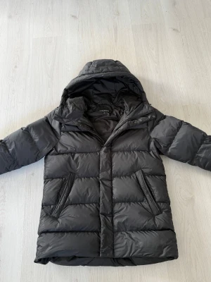 Svart dunjacka med huva från Lindbergh - Säljer en svart dunjacka från J.Lindeberg med huva och dragkedja. Jackan har två snedställda fickor framtill och breda, quiltade paneler som ger en schysst look. Perfekt för kalla vinterdagar och riktigt skön att bära. Passar M/L. Pris går absolut att diskutera! Hör av er!