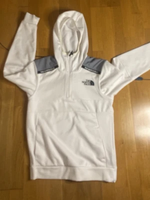 Vit hoodie från The North Face - Snygg vit hoodie från The North Face med halv dragkedja och grå detaljer på axlarna. Tillverkad i ett strukturerat, tekniskt material som känns sportigt och modernt. Stor logga på bröstet och huva för extra stilpoäng.