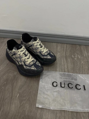 Gucci sneakers med monogrammönster - Säljer ett par snygga Gucci sneakers med klassiskt blått GG-monogram på beige tyg. Skorna har vita snören, mörkblå detaljer och en chunky sula. Perfekta för dig som vill sticka ut med exklusiv design och streetwear-vibe.