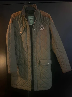 Olivgrön quiltad cardiganjacka Lexington - Snygg olivgrön quiltad cardiganjacka från Lexington med bruna axelpartier och broderad logga på bröstet. Jackan har tryckknappar, fickor med lock och en längre passform. Perfekt för dig som gillar klassisk och sportig stil.